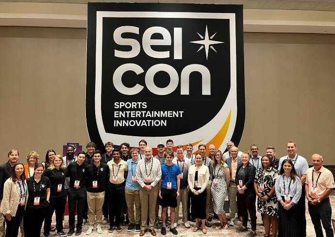 Group of people posing in front of a banner that reads 'SEI CON SPORTS ENTERTAINMENT INNOVATION,' with many wearing name badges and dressed in business casual attire.