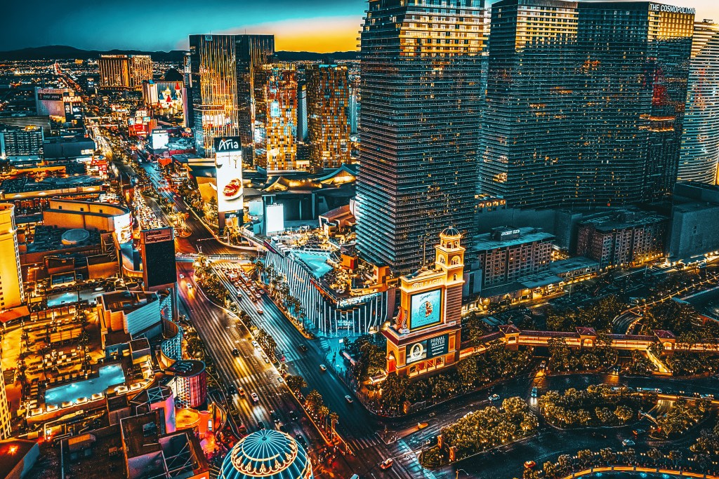 Aerial view of the Las Vegas Strip at dusk, with numerous brightly lit hotels, casinos, and billboards, including the Bellagio and Aria.