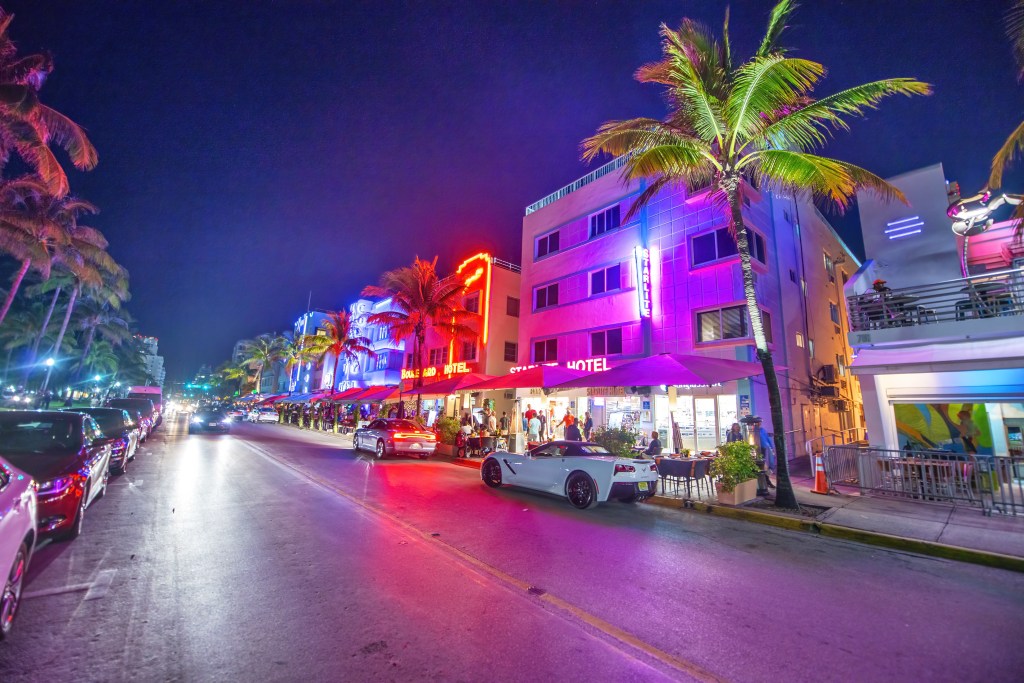 Ocean Boulevard at night with palm trees and illuminated buildings housing restaurants with outdoor seating, cars parked and driving.